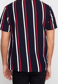 Košile s krátkým rukávem v námořnické modři, s výraznými vertikálními pruhy v červené a bílé barvě, s kulatým výstřihem a volným střihem.