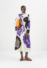 Knit, long-sleeve dress in cream with large purple and brown floral patterns. Model holds an orange clutch, wearing brown strappy sandals.