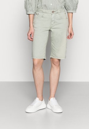 Personne portant un short beige clair arrivant aux genoux, une chemise à manches bouffantes rayée et des baskets blanches, debout devant un fond uni.