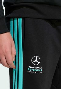 Un pantalon de survêtement noir présente trois bandes verticales turquoise sur le côté, avec un logo Mercedes blanc et un texte sur la partie inférieure de la jambe.