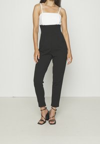 Femme portant un haut sans manches blanc et un pantalon noir taille haute, associé à des sandales à talons à lanières noires, se tenant devant un mur uni.