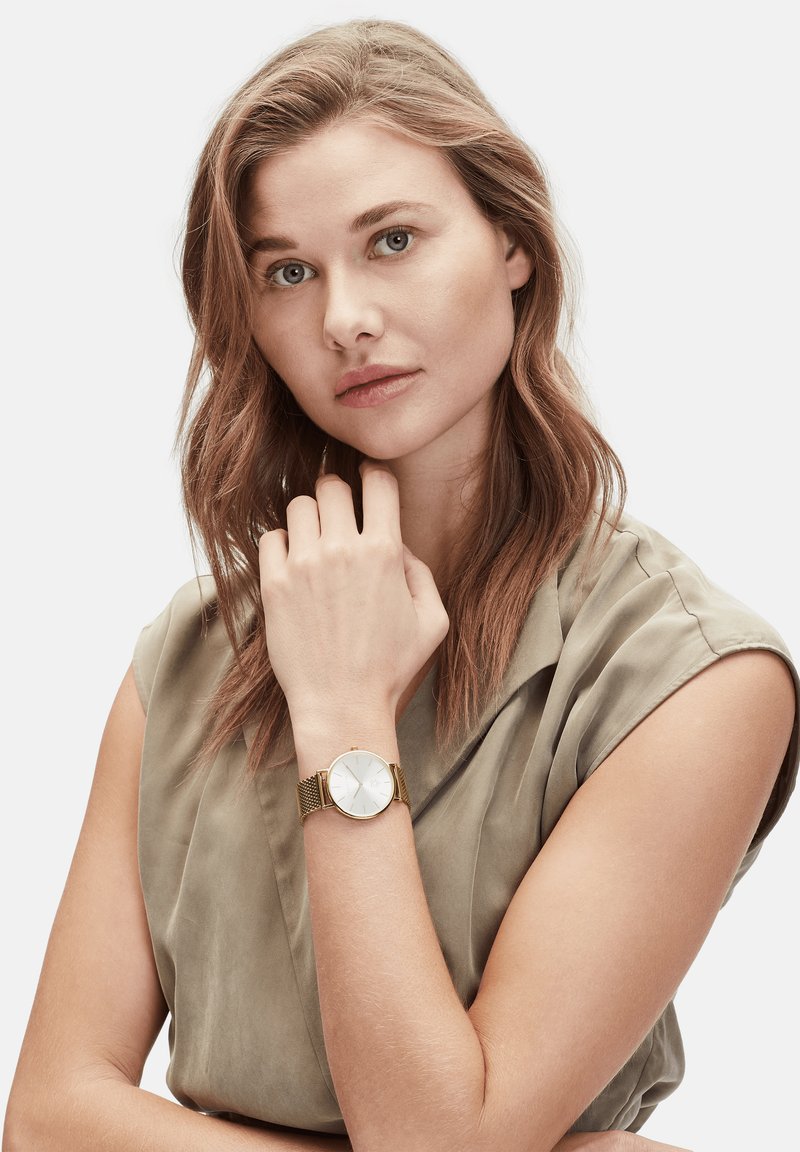 Montre-bracelet en maille dorée avec un cadran rond blanc et un affichage minimaliste. Le modèle porte un haut vert clair, posant avec la main sur le menton.