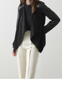 Veste noire avec un devant drapé, matériaux en daim et cuir combinés, manches longues et col noir côtelé, portée sur un pantalon blanc.