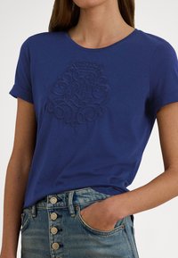 Γυναίκα που φοράει μπλε κοντομάνικη μπλούζα με ανάγλυφο σχέδιο βασιλικού οικόσημου, βεβιδωμένη σε ψηλόμεσα μπλε τζιν με κουμπιά.