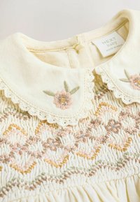 Robe pour bébé de couleur crème avec un col en dentelle, ornée de broderies florales délicates dans des teintes pastel de rose et de vert, ainsi que de détails froncés.