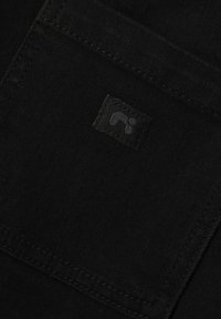 Tela negra con una textura suave, destacada por una pequeña etiqueta negra con un logotipo cosido en un bolsillo, acentuada por sutiles detalles de costura.