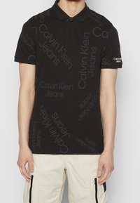 Svart polo skjorta med upprepande grå "Calvin Klein Jeans" textmönster och vit "Calvin Klein Jeans" logotyp på vänster ärm, bärs tillsammans med beige byxor.