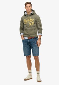 Olivengrøn hættebluse med gul tekst, denimshorts, beige sneakers og stribede skjorteærmer. Har en forlommer og et brunt bælte.