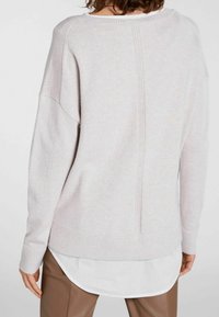 Hellgrauer Pullover mit lockerer Passform, gerippten Bündchen und einer zentralen Naht. Über einem weißen Hemd mit abgerundetem Saum getragen. Weiche Textur.