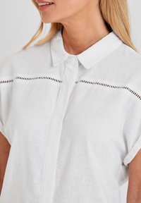 White linen shirt featuring a button-up front, collar, short rolled sleeves, and a horizontal cut-out detail across the chest.