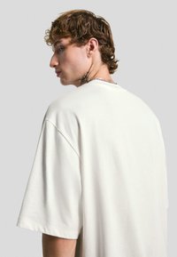 Mladenič z kodrastimi rjavimi lasmi nosi prevelik preprost bel T-shirt, ki razkriva tetovažo na vratu in srebrno verigo, ter gleda na stran.