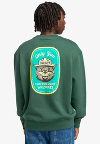 Felpa verde con una grafica di un orso che indossa un cappello da ranger. Il testo dice: "Solo tu puoi prevenire gli incendi boschivi." Polsini e orlo a coste.