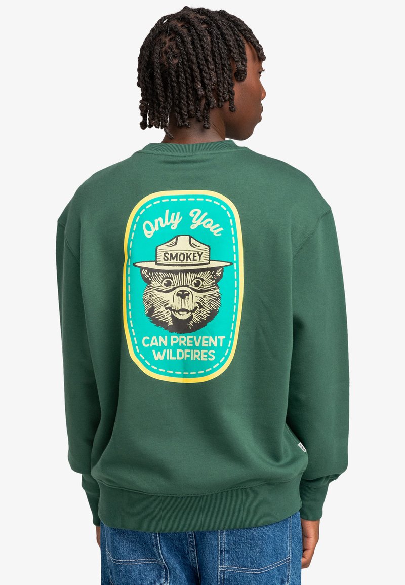 Felpa verde con una grafica di un orso che indossa un cappello da ranger. Il testo dice: "Solo tu puoi prevenire gli incendi boschivi." Polsini e orlo a coste.