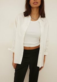 Bílá košile na knoflíky z lehkého materiálu, nošená rozepnutá přes bílý žebrovaný crop top. Tmavé kalhoty s vysokým pasem, masivný stříbrný řetězový náhrdelník.