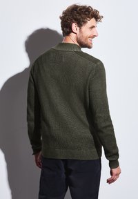 Pletený svetr v tmavě zelené barvě s texturovaným vzorem, žebrovaným límcem, manžetami a spodním lemem; model stojící bokem, předvádějící design na zadní straně.