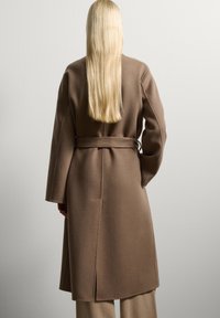 Cappotto lungo in lana marrone con vita cinturata, maniche ampie e taglio dritto, con fenditure sui lati per facilitare il movimento.