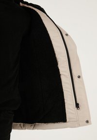 Beige jacka med en mjuk svart fleeceinredning. Har en slät utsida, blixtlås och silverknappar. Texturerad tyg kontrasterar med foder.
