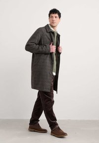 Hnedý károvaný kabát s veľkým golierom, nosený cez zelený sveter a hnedé kordové nohavice; hnedé topánky dotvárajú celkový outfit.