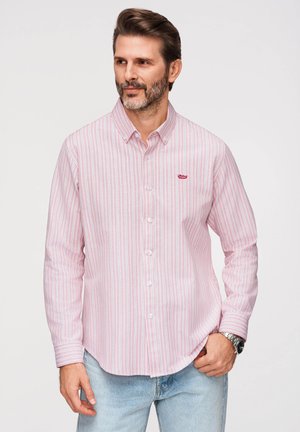 Mężczyzna w jasnoróżowej i białej koszuli w paski zapinanej na guziki oraz jasnoniebieskich dżinsach, z zegarkiem na lewym nadgarstku, stojący na jednolitym tle.