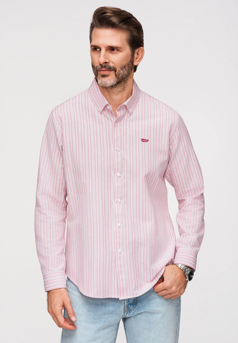 Homme portant une chemise à rayures rose clair et blanche avec boutons et un jean bleu clair, avec une montre au poignet gauche, debout devant un fond uni.