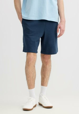 Hombre con pantalones cortos azul marino, camisa azul clara, calcetines blancos y zapatillas blancas, de pie frente a un fondo claro y liso.