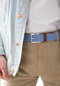 Woven blauer Gürtel mit braunen Lederakzenten, silberner quadratischer Schnalle, getragen über einer hellen Hose und einem weißen Hemd unter einer hellen Jeansjacke.