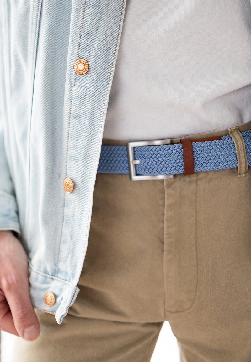 Woven blauer Gürtel mit braunen Lederakzenten, silberner quadratischer Schnalle, getragen über einer hellen Hose und einem weißen Hemd unter einer hellen Jeansjacke.