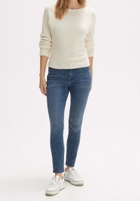 Pull à côtes crème, jean bleu ajusté et baskets blanches avec des accents verts. Texture douce et silhouette élancée. Tenue décontractée.
