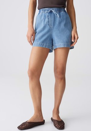Woman wearing light blue denim shorts with elastic waistband and drawstring, paired with dark woven slip-on shoes.