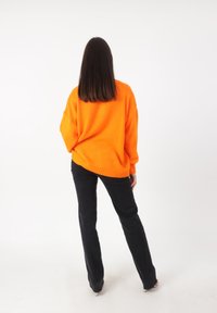 Pullover arancione brillante di grandi dimensioni con polsini a coste, abbinato a pantaloni neri aderenti. Lo sfondo semplice mette in risalto i colori vivaci.