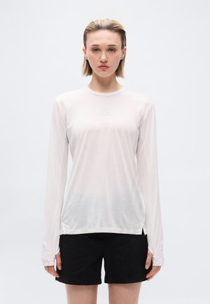 Woman with short blonde hair wearing a sheer white long-sleeve top and black shorts, standing against a plain light background.