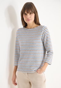 Femme aux cheveux bruns lisses portant un pull à manches longues rayé blanc, bleu et marron, et un pantalon beige, debout avec une main dans la poche.