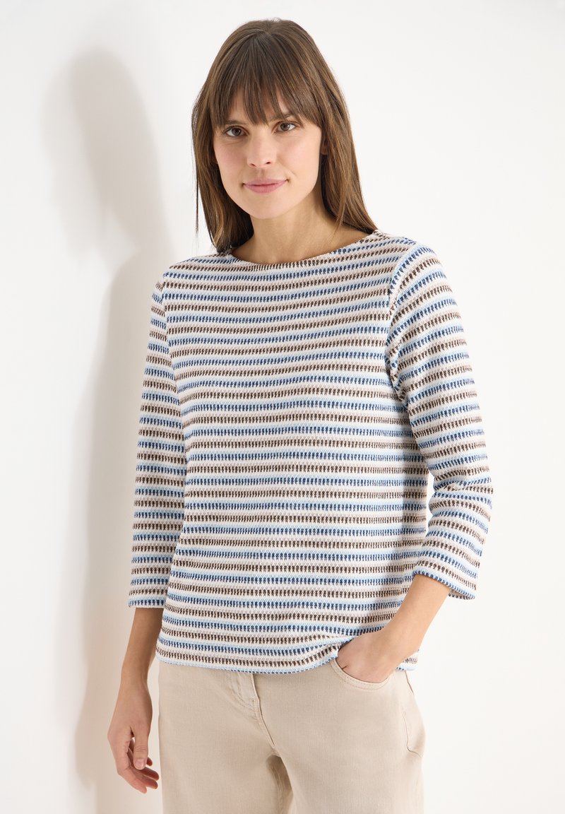 Femme aux cheveux bruns lisses portant un pull à manches longues rayé blanc, bleu et marron, et un pantalon beige, debout avec une main dans la poche.