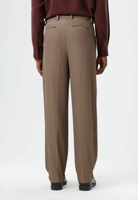 Pantalon beige rayé à coupe ample, avec des plis, deux poches arrière et une ceinture avec passants. Porté avec une chemise bordeaux à manches longues.