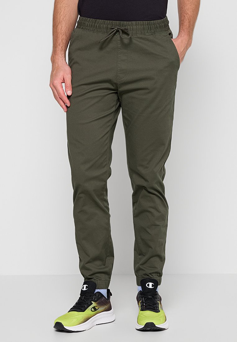 Pantalons de jogging vert olive avec une taille élastique et un cordon de serrage. Dispose de poches latérales et de chevilles fuselées. Tissu en coton lisse.