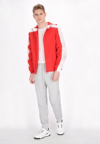 Rød og hvid zip-up jakke med hætte, parret med grå sweatpants og hvide sneakers. Jakken har elastiske manchetter og et sporty design.