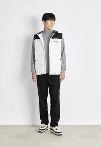 Balti pūkuoti liemenė su juodais peties akcentais, pilkas golfas, juodos kelnės ir kreminiai bei juodi sportbačiai, stovintys priešais šviesų foną.