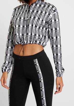 Veste Adidas noir et blanc courte avec des bandes logo, associée à des leggings noirs avec des bandes logo Adidas sur les côtés.