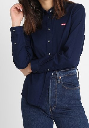 Kobieta w granatowej koszuli Levi's na guziki, wpuszczonej do jeansów z wysokim stanem w ciemnoniebieskim kolorze, z jedną ręką skrzyżowaną i dłonią blisko twarzy.