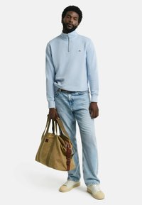 Hellblauer Viertelreißverschluss-Pullover, beige Jeans, cremefarbene Sneakers. Modell hält eine beigefarbene Sporttasche mit braunen Lederdetails und -trägern.