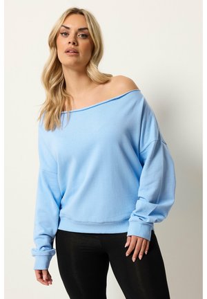 Lyshåret kvinde iført en lyseblå off-shoulder sweatshirt og sorte leggings, poserer mod en ensfarvet hvid baggrund.