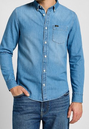 Mand iført en lyseblå denimskjorte med knapper og brystlomme og mørkeblå denimjeans, hånd i lommen på den ene side.
