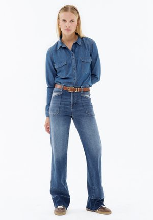 Denim button-up shirt with long sleeves, two chest pockets. High-waisted wide-leg jeans in light blue with a brown belt and sneakers.