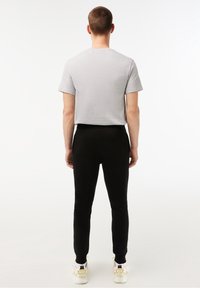 Mand stående med ryggen til, iført en lysegrå kortærmet t-shirt, sorte bukser og hvide sneakers med gule detaljer.