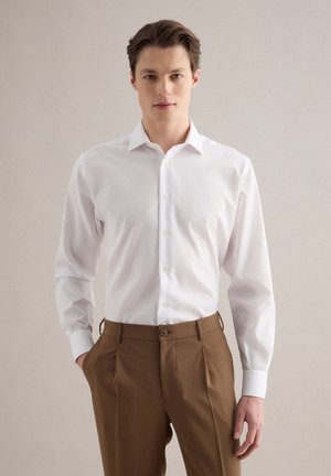 Junger Mann trägt ein weißes Hemd mit Knöpfen und braune plissierte Hosen, steht vor einem schlichten neutralen Hintergrund.