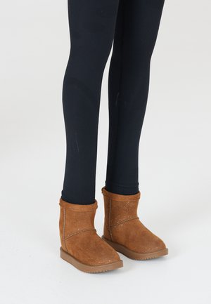Stivaletti alla caviglia in suede marrone con una consistenza morbida, cuciture in rilievo e suola in gomma piatta, abbinati a leggings neri aderenti.