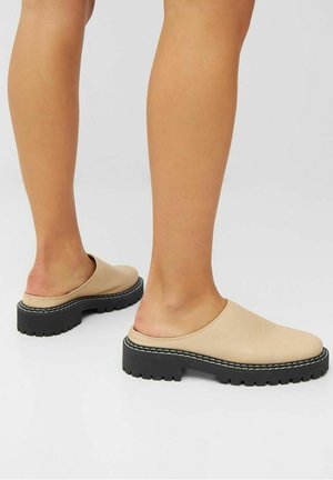 Beine, die beige Slip-On-Schuhe mit dicken schwarzen Sohlen und weißen Nähten tragen, stehen auf einer hellgrauen Oberfläche.