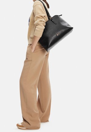 Frau trägt einen beigen Pullover und Hosen mit weitem Bein, trägt eine große schwarze Einkaufstasche über der Schulter vor weißem Hintergrund.