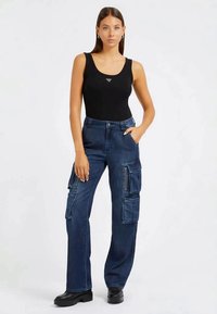 Visoke modre jeans hlače s številnimi stranskimi žepi, ohlapen kroj, zadrže in subtilno izbledi pranje. Nosi se z črno majico brez rokavov.