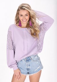 Lavendelkleurige trui met kantdetails op de mouwen, ronde halslijn en lange mouwen, gecombineerd met versleten denim shorts.